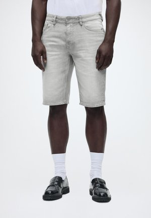 Muž oblečený v svetlosivých džínsových šortkách, bielych ponožkách, čiernych kožených mokasínach a bielom tričku, stojaci na jednofarebnom pozadí.