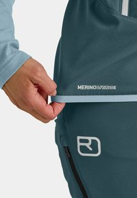 Merinowoll-Mischgewebe in blaugrau. Hat eine hellblaue Einfassung, eine Reißverschlusstasche und weiße Markenbeschriftung. Die Textur wirkt glatt und atmungsaktiv.