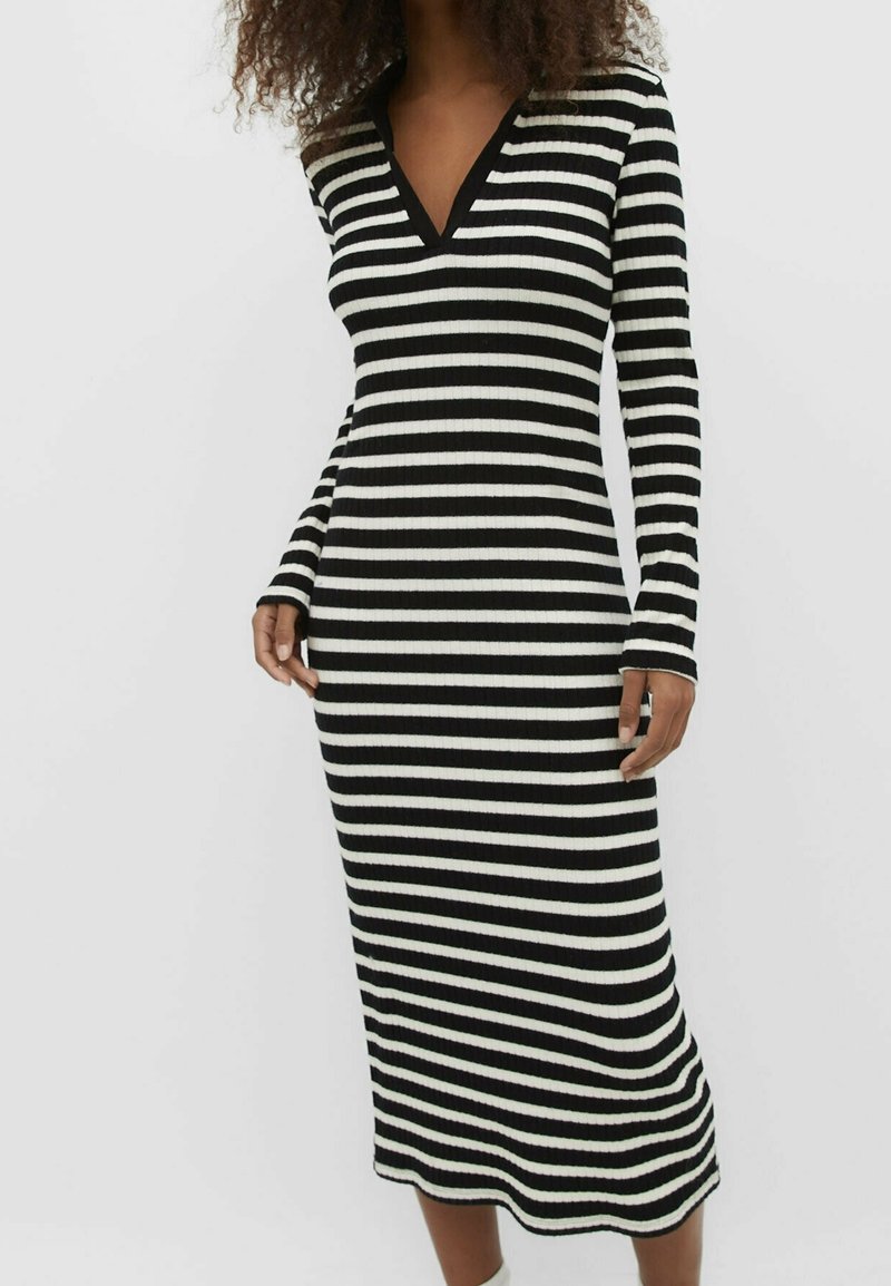 Femme portant une robe midi à manches longues rayée horizontalement noir et blanc avec un col en V, sur un fond clair uni.