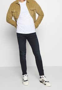Tan corduroy shirt over a white t-shirt, dark slim-fit jeans, and white sneakers with black, red, and blue accents.