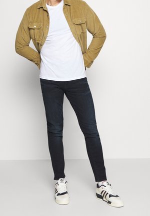 Tan corduroy shirt over a white t-shirt, dark slim-fit jeans, and white sneakers with black, red, and blue accents.