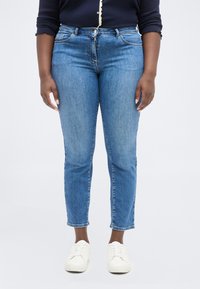 Άτομο που φοράει μπλε τζιν, λευκά αθλητικά παπούτσια και μπλε μπλουζάκι μακρυμάνικο με κυματιστές άκρες στα μανίκια και κουμπιά.