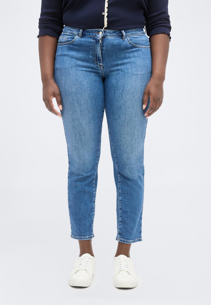 Άτομο που φοράει μπλε τζιν, λευκά αθλητικά παπούτσια και μπλε μπλουζάκι μακρυμάνικο με κυματιστές άκρες στα μανίκια και κουμπιά.