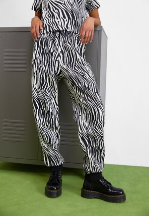 Personne portant un pantalon imprimé zèbre et un haut assorti, appuyée contre des casiers gris, avec des bottes plateformes noires épaisses sur un sol vert.