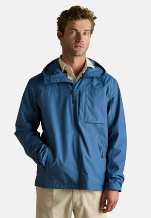 Mann in einer blauen kapuzenjacke mit Reißverschluss und Brusttasche, trägt beige Cordhosen, steht vor einem einfarbigen Hintergrund.