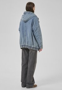 Giacca di jeans azzurra chiara con cappuccio, vestibilità oversize, orlo elastico e spalle dropping. Abbinata a pantaloni larghi grigio scuro.