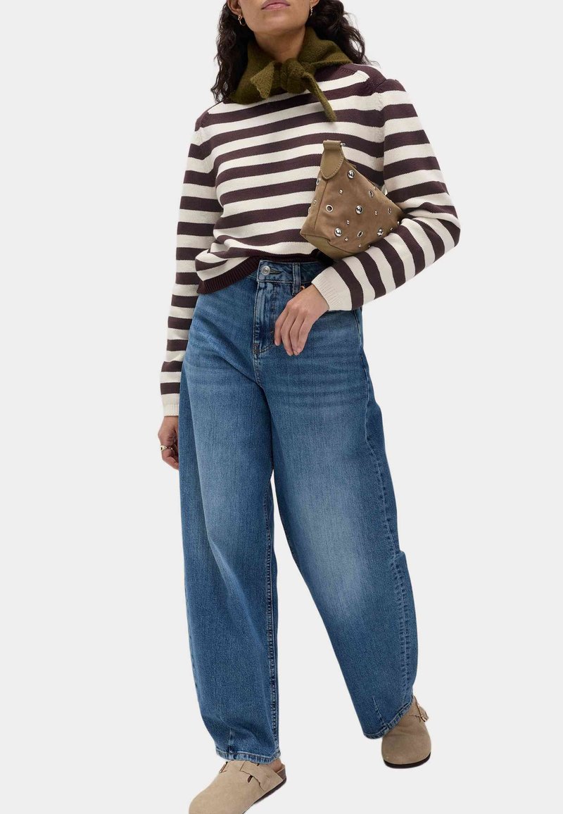 Femme portant un pull rayé marron et blanc, un jean bleu à jambes larges, des chaussures beiges, et tenant un sac à main marron clouté sous le bras.