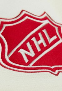 Rotes gesticktes NHL-Logo auf einem cremefarbenen Textilhintergrund, das eine schildförmige Form zeigt, mit weißen Akzentnähten um das Emblem.