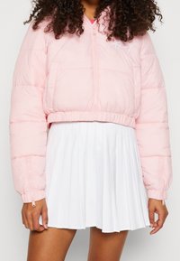Kvinna i en ljusrosa kort puffjacka med dragkedja på ärmarna och en vit plisserad kjol, stående mot en neutral bakgrund.