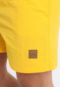 Shorts de bain jaunes en tissu léger. Présentent une patch carrée marron avec la marque "URBAN CLASSICS". Surface texturée, longueur courte.
