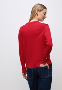 Roter Strickpullover mit langen Ärmeln, runder Ausschnitt und Knopf-Details an den Manschetten. Kombiniert mit dunkelblauen Jeans.