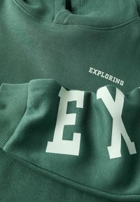 Felpa verde con testo bianco che leggono "EXPLORING" e "EX." Realizzata in tessuto morbido, con polsini a coste e cappuccio.