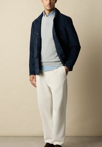 Marineblauw jasje over een grijze trui, lichtblauw shirt eronder. Witte geribbelde broek en koffiebruine schoenen. Soepel textiel met gelaagd ontwerp.