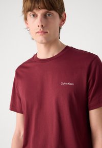 Burgundská bavlnená tričko s krátkymi rukávmi, okrúhlym výstrihom a bielym logom "Calvin Klein" na ľavom prsiach. Hladká štruktúra.