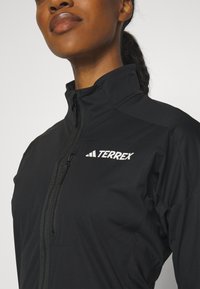 Schwarze Zip-Jacke aus leichtem Stoff, mit hohem Kragen und weißem "TERREX"-Logo auf der Brust. Geschmeidige Textur mit körperbetontem Design.