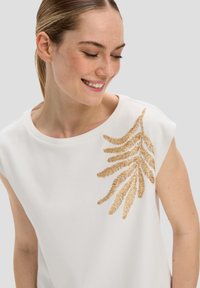 T-shirt blanc à manches courtes présentant un design de fougère brodé en or sur l'épaule gauche, avec une texture de tissu lisse.