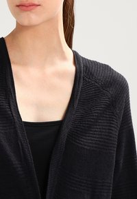 Czarny kardigan o teksturowanym wzorze, z dekoltem w kształcie litery V i otwartym przodem. Tkanina ma miękką strukturę i delikatne poziome paski.