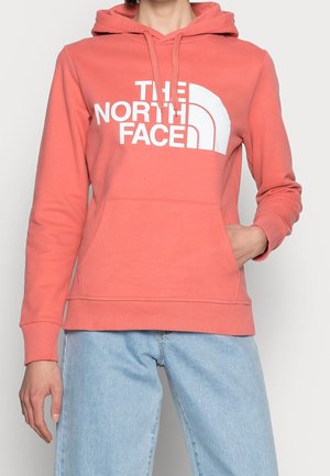 Person wearing coral pink The North Face hoodie and light blue jeans, with one hand in the hoodie pocket, against a plain background.