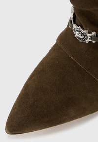 Stivale alla caviglia in suede marrone con punta affusolata e accentuazione decorativa in metallo. La texture liscia con una finitura morbida esalta il design.