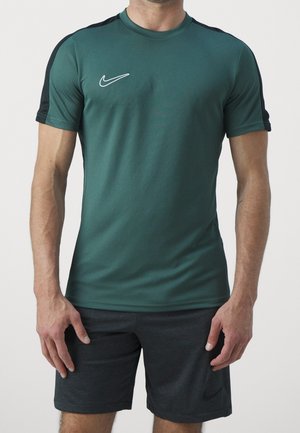 Homme portant un t-shirt de sport Nike à manches courtes vert foncé avec des bandes noires sur les épaules et un short Nike gris foncé, debout devant un fond uni.