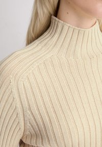 Beige geribbelde gebreide coltrui met een gestructureerde afwerking. Kenmerkt zich door een hoge halslijn en licht gestructureerde schouders.