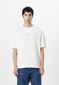 Weißes Baumwoll-T-Shirt mit lockerem Schnitt, Rundhalsausschnitt und kurzen Ärmeln. Auf der Vorderseite sind kleine blaue Grafiken zu sehen. Denim-Jeans sind sichtbar.
