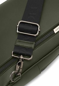 Olivengrüne synthetische Tasche mit glatter Textur, ausgestattet mit einem gepolsterten schwarzen Tragegurt, silbernen Beschlägen und einem Reißverschlussdetail.