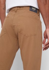 Beige Hosen aus Baumwollmischung, mit zwei Gesäßtaschen und einem gewebten Etikett am Bund. Die Textur ist glatt mit dezenten Nähten.