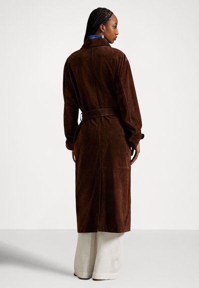 Žena s vrkočmi, oblečená v dlhom hnedom semišovom kabáte s opaskom, modrej košeli, bielych širokých nohaviciach a visiace náušnice, otočená bokom.