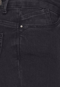 Jeans de denim preto com uma textura suave, design clássico de cinco bolsos e um pequeno detalhe em metal na parte frontal.