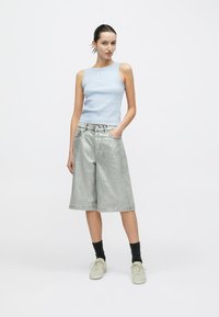 Γαλάζιο αμάνικο τοπ, ασημένια μεταλλική βερμούδα με φαρδιά πόδια και ανοιχτόχρωμα ουδέτερα αθλητικά παπούτσια. Το μοντέλο στέκεται μπροστά σε απλό φόντο.