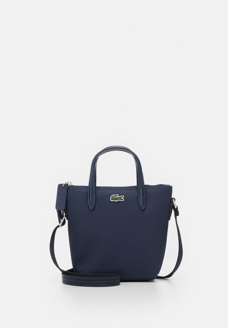 Sac à main bleu marine avec un design structuré, une texture lisse, des poignées courtes et une bandoulière amovible. Logo Lacoste sur le devant.
