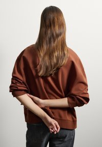 Felpa marrone oversize con una vestibilità rilassata, maniche arricciate e polsini a coste, abbinata a jeans di denim scuri. Capelli lunghi e lisci in vista.