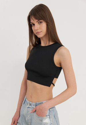 Schwarzes, geripptes Crop-Top mit rundem Ausschnitt und ärmellosem Design. Mit Cut-Out-Details an den Seiten. Kombiniert mit glänzenden hellblauen Jeans.