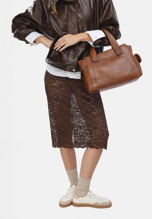 Personne portant une jupe en dentelle marron, des baskets blanches avec des chaussettes beiges, une veste en cuir marron foncé, tenant un sac à main en cuir marron.