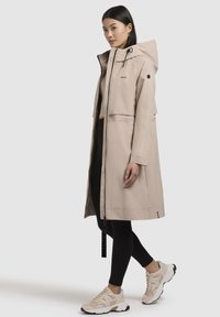 Beige regenjas met capuchon en ritsluiting, met een losse pasvorm en zijzakken. Gedragen over zwarte leggings en lichte sneakers.