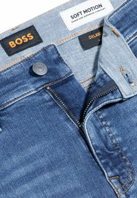 Vaqueros de mezclilla en un tono azul medio con costuras naranjas. Presentan un cierre con cremallera, broche y etiquetas de BOSS y SOFT MOTION.