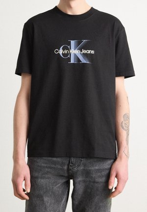 Personne portant un t-shirt noir Calvin Klein Jeans avec un grand logo CK bleu et blanc et un jean noir délavé. Tatouage visible sur l'avant-bras droit.