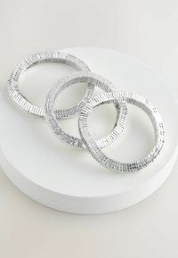 Sæt med tre sølvfarvede metalarmbånd med et tekstureret design, der har en rund form og varierende tykkelser, præsenteret på en hvid pedestal.