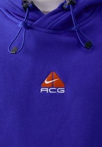Felpa viola con cappuccio e cordini, caratterizzata da un logo ACG di Nike ricamato in arancione e bianco. Realizzata in un tessuto morbido e resistente con una finitura testurizzata.