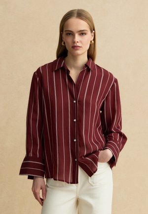 Vrouw draagt een bordeauxrode blouse met knopen en witte verticale strepen, gecombineerd met crème broek, staand voor een beige achtergrond.