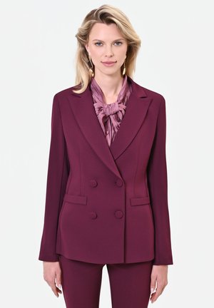 Donna con capelli biondi indossa un blazer doppiopetto viola scuro e pantaloni abbinati con una camicetta viola chiaro annodata al collo con un fiocco.