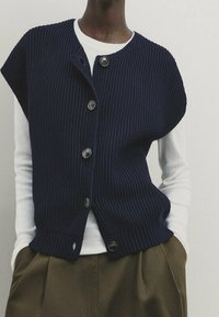 Laivastonsininen ribbineulottu liivi, jossa on lyhyet hihat, viisi suurta nappia ja rento istuvuus, kerrostettuna valkoisen pitkähihaisen paidan päälle.