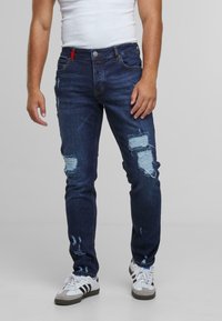 Herre mørkeblå denim jeans med slitt detaljer, som har flere rifter og en slank passform. Brukt med hvite joggesko og en tettsittende hvit tanktop.