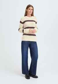 Gebreide trui in crèmewit met bruine horizontale strepen, ronde hals, ribbeltextuur; gecombineerd met een wijde jeans in het blauw en zwarte schoenen.