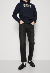 Persona vestida con una sudadera azul marino con el texto "GANT", jeans negros, calcetines blancos y mocasines negros, de pie con las manos en los bolsillos frente a un fondo sencillo.