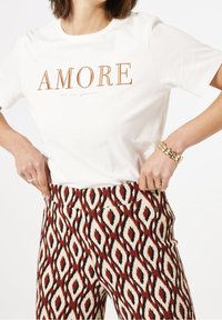 Biała koszulka z napisem "AMORE" w miedzianej haftowanej nitce, połączona z wzorzystymi spodniami z diamentowym wzorem w kolorach czerwonym, czarnym i kremowym.