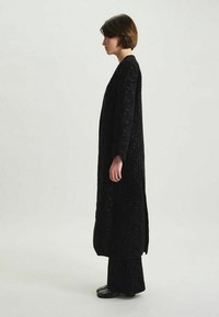Long manteau noir en tissu texturé, avec des manches larges et des fentes sur les côtés; porté sur un pantalon noir assorti et des chaussures noires, présenté de côté.