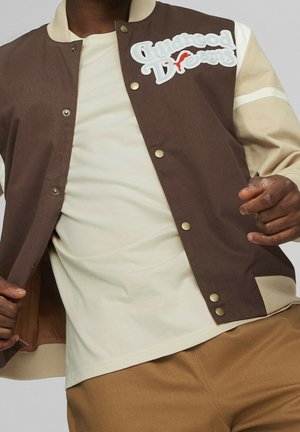 Hombre vestido con una chaqueta de béisbol marrón y beige con un parche que dice "Sueños de infancia", camisa crema y pantalones marrones, posando con los brazos doblados.