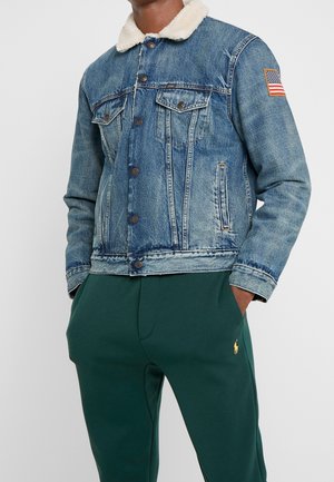 Mand iført blå denimjakke med hvidt shearling-krave og amerikansk flagpatch på ærmet, kombineret med mørkegrønne bukser med gul logo.